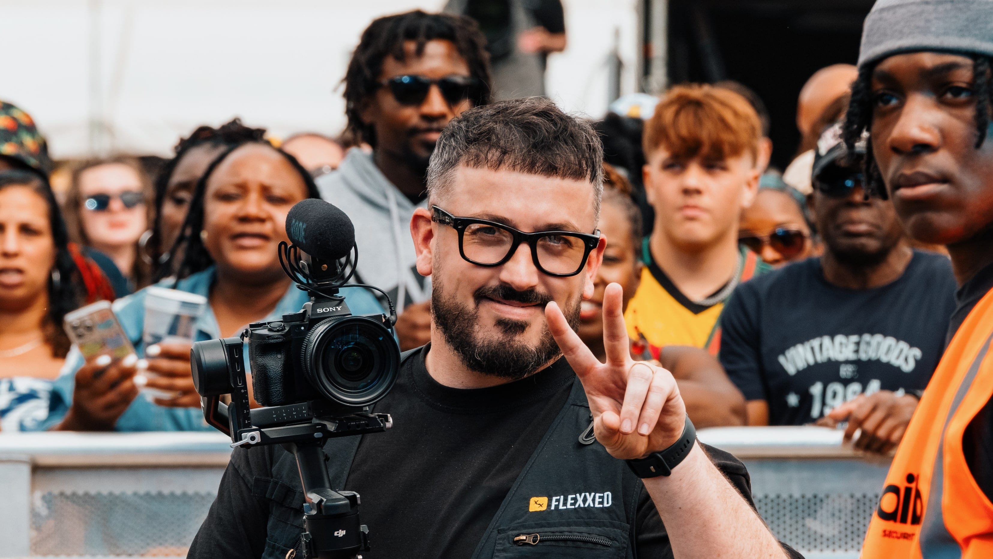 A Flexxed camera operator holding a gimbal rig at the front of a festival crowd, the bar and the barrier behind him