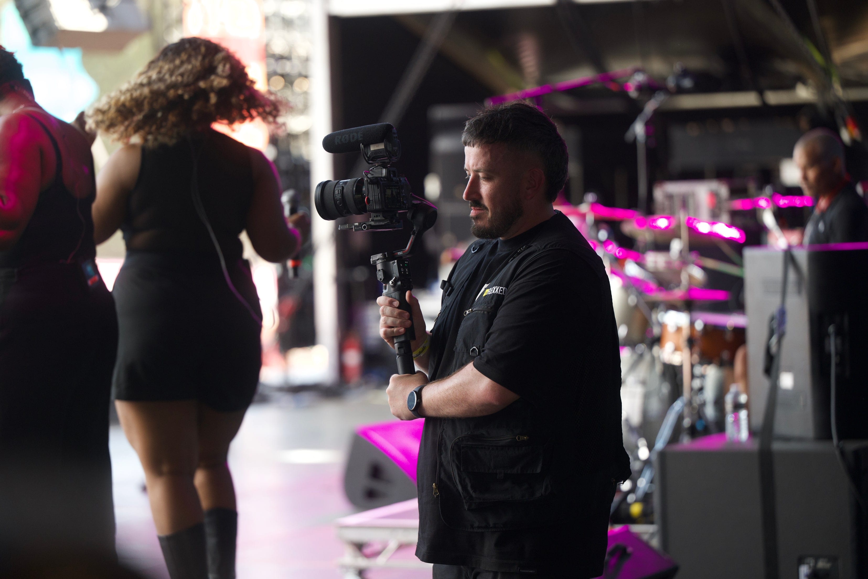 A Flexxed operator filming on a gimbal at the side of the Boogietown stage, drum kit and pink stage light behind