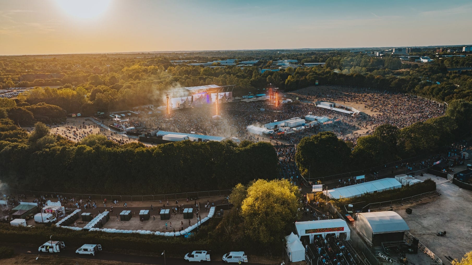 Golden-hour aerial of a lit festival stage and the crowd in front of it, treeline and site beyond