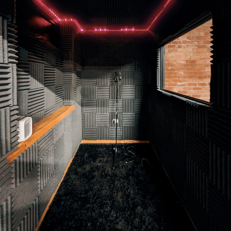 Vocal booth lined with acoustic foam, a microphone and a window to the control room