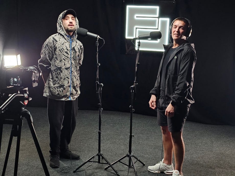 Two performers at microphone stands on the studio floor, the lit Flexxed sign behind them and a camera rig in the foreground