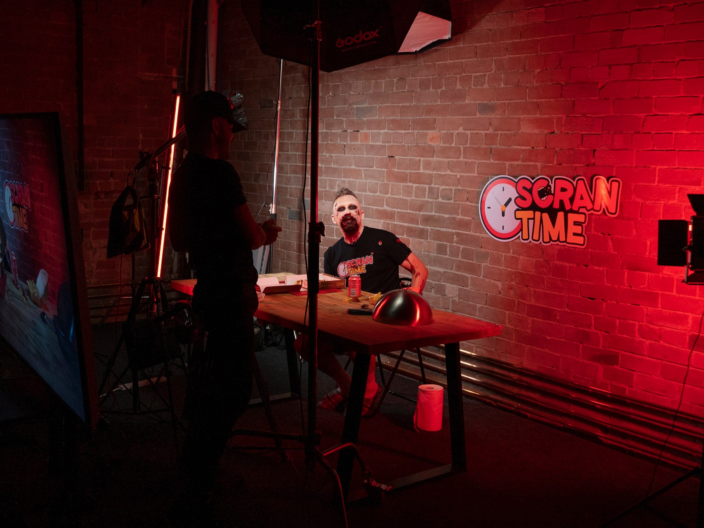 Presenter at the desk under a red neon Scran Time sign, camera in the foreground